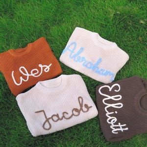 K&ouml;nnte beinhalten: Vier Strickpullover in den Farben Rost, Creme und Dunkelbraun. Jeder Pullover hat einen Rundhalsausschnitt und einen auf der Vorderseite mit kontrastierendem Garn gestickten Namen: Wes in Wei&szlig;, Abraham in Hellblau, Jacob in Braun und Elliott in Wei&szlig;.