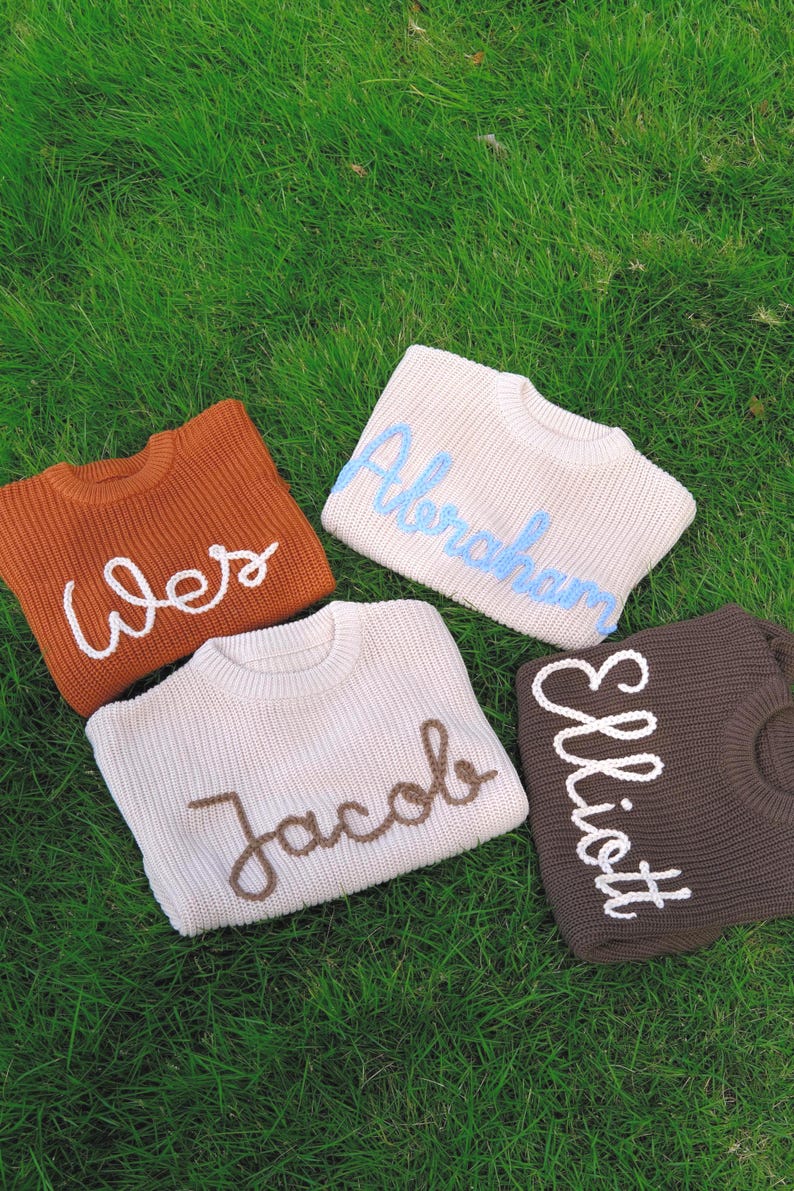 K&ouml;nnte beinhalten: Vier Strickpullover in den Farben Rost, Creme und Dunkelbraun. Jeder Pullover hat einen Rundhalsausschnitt und einen auf der Vorderseite mit kontrastierendem Garn gestickten Namen: Wes in Wei&szlig;, Abraham in Hellblau, Jacob in Braun und Elliott in Wei&szlig;.