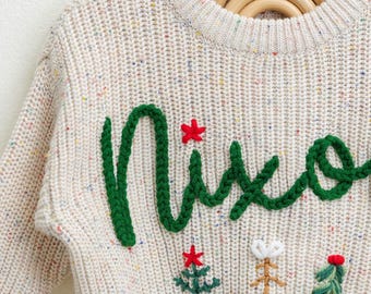 May include: A white knitted sweater with a green embroidered "Nice" and colorful embroidered Christmas trees.
