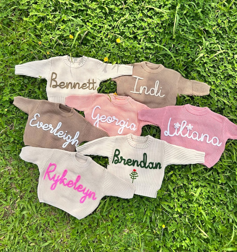 K&ouml;nnte beinhalten: Sieben lang&auml;rmelige Strickpullover in verschiedenen Farben wie Creme, Beige, Braun und Rosa. Jeder Pullover ist auf der Vorderseite mit einem Namen in Schreibschrift bestickt: Bennett, Indi, Everleigh, Georgia, Liliana, Rykeleyn und Brendan mit einem Baum.