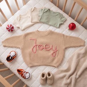 K&ouml;nnte beinhalten: Ein beigefarbener, lang&auml;rmeliger Strickpullover mit dem in rosa Garn aufgestickten Namen Joey auf der Vorderseite, dazu drei kurz&auml;rmelige Bodys in Creme und Salbeigr&uuml;n, zwei orange-wei&szlig;e Strick-Sneaker und ein Paar beigefarbene Stricksch&uuml;hchen.