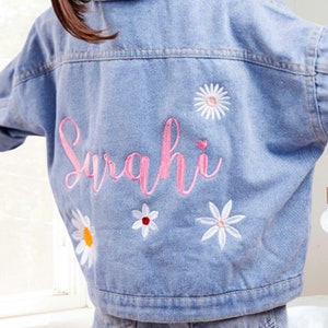 May include: A light blue denim jacket with the name "Sarahi" embroidered in pink on the back. The jacket is decorated with white embroidered flowers.