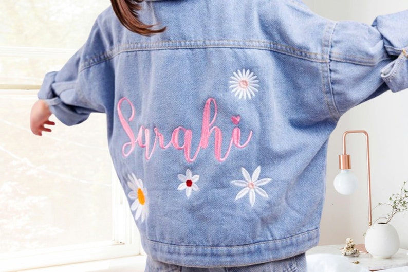 May include: A light blue denim jacket with the name "Sarahi" embroidered in pink on the back. The jacket is decorated with white embroidered flowers.