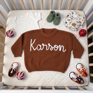 K&ouml;nnte beinhalten: Ein rostfarbener Strickpullover mit dem in wei&szlig;er Schreibschrift gestickten Namen Karson, dazu ein wei&szlig;er Body, olivgr&uuml;ne Stricksch&uuml;hchen, eine gemusterte Stoffwindel sowie zwei Paar orange-wei&szlig;e Turnschuhe mit roten Weihnachtskugeln.