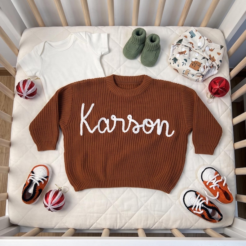 K&ouml;nnte beinhalten: Ein rostfarbener Strickpullover mit dem in wei&szlig;er Schreibschrift gestickten Namen Karson, dazu ein wei&szlig;er Body, olivgr&uuml;ne Stricksch&uuml;hchen, eine gemusterte Stoffwindel sowie zwei Paar orange-wei&szlig;e Turnschuhe mit roten Weihnachtskugeln.