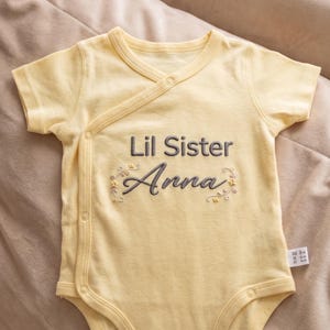 Op de afbeelding: Een lichtgele babybody met korte mouwen en een wikkelvoorkant. De tekst "Lil Sister Anna" is geborduurd in grijs schrift met bloemaccenten. De body heeft drukknoopsluitingen en is gemaakt van zachte stof.