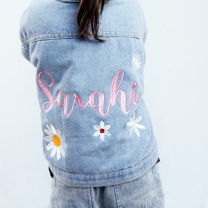 May include: A light blue denim jacket with the name "Sarahi" embroidered in pink on the back. The jacket is decorated with white daisy flowers.