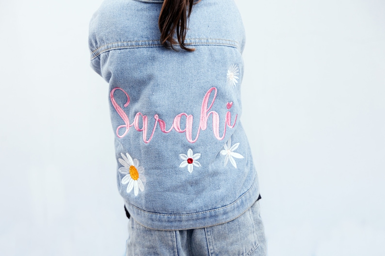 May include: A light blue denim jacket with the name "Sarahi" embroidered in pink on the back. The jacket is decorated with white daisy flowers.