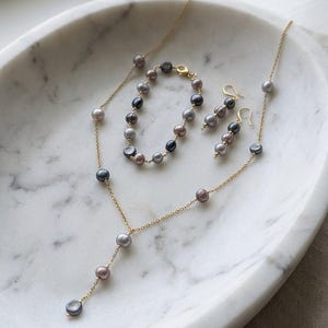 Op de afbeelding: Een sieradenset met een gouden ketting, armband en oorbellen. De set bevat grijze, zwarte en mauvekleurige parels. De sieraden worden gepresenteerd op een wit marmeren dienblad.