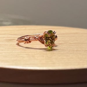 May include: A handmade copper wire ring with a heart-shaped, light green gemstone. The ring is set on a wooden surface, showcasing the intricate wire wrapping around the gemstone. The ring's design is delicate and unique.