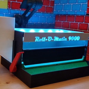 May include: A black and white Roll-O-Matic 9000 machine with a red lever and a white box. The machine has a green base and is illuminated with blue lights. The text "Roll-O-Matic 9000" is displayed on the front.