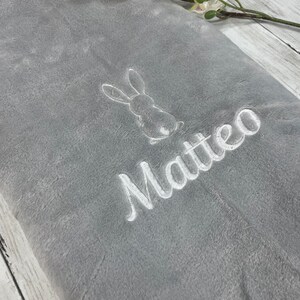 Pode incluir: Um cobertor de pel&uacute;cia cinza com um coelho bordado em branco e o nome "Matteo" nele.