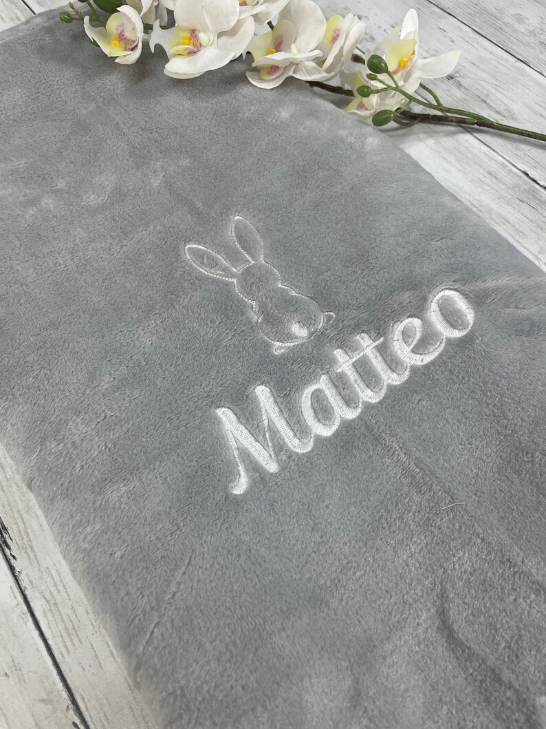 Pode incluir: Um cobertor de pel&uacute;cia cinza com um coelho bordado em branco e o nome "Matteo" nele.