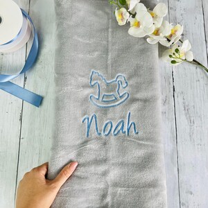 Pode incluir: Um cobertor de beb&ecirc; de pel&uacute;cia cinza com bordado azul de um cavalo de balan&ccedil;o e o nome "Noah".