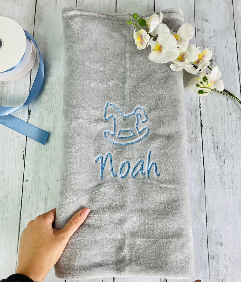 Pode incluir: Um cobertor de beb&ecirc; de pel&uacute;cia cinza com bordado azul de um cavalo de balan&ccedil;o e o nome "Noah".