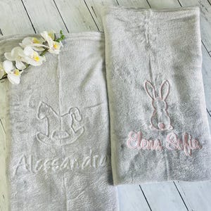 Pode incluir: Duas mantas de beb&eacute; cinzas com nomes e desenhos bordados. Uma manta apresenta um cavalo de baloi&ccedil;o e o nome Alessandro. A outra manta apresenta um coelho e os nomes Elena Sofia.