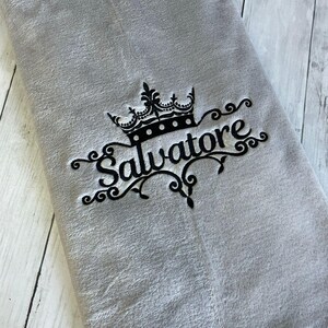 Pode incluir: Uma manta de l&atilde; cinza com um desenho bordado preto de uma coroa e a palavra "Salvatore".