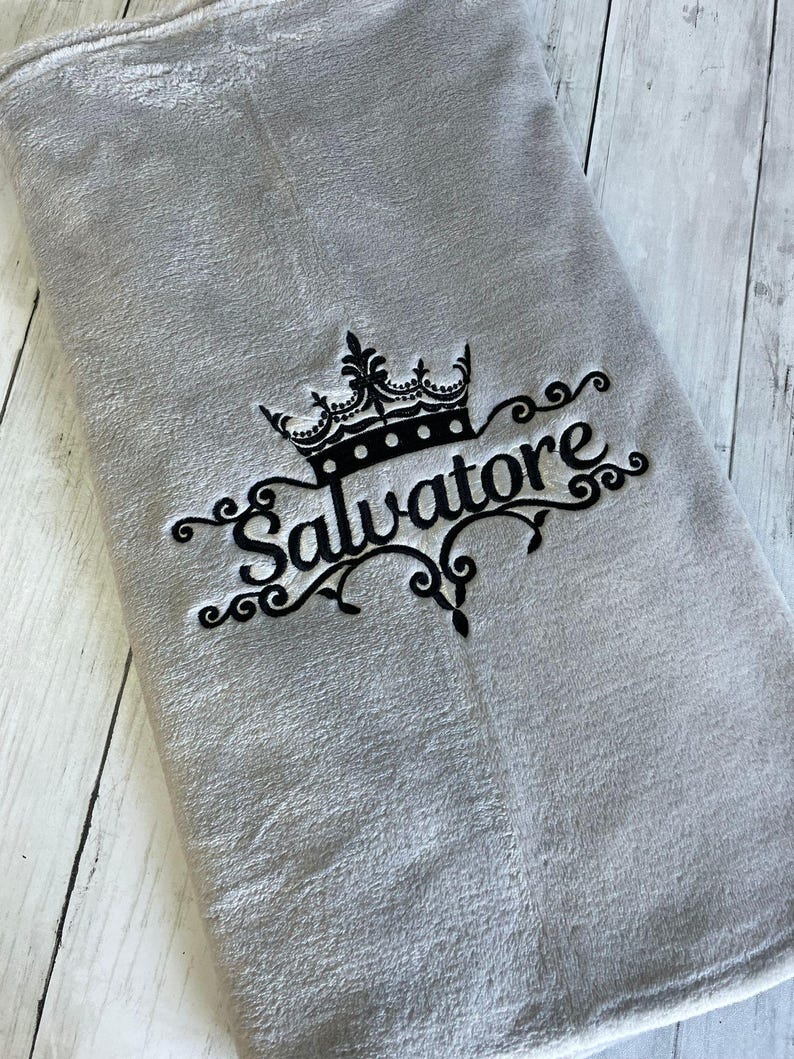 Pode incluir: Uma manta de l&atilde; cinza com um desenho bordado preto de uma coroa e a palavra "Salvatore".