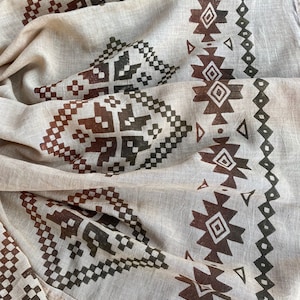 May include: A beige cotton scarf with a brown and black geometric pattern. The pattern is a repeating design of squares and triangles.