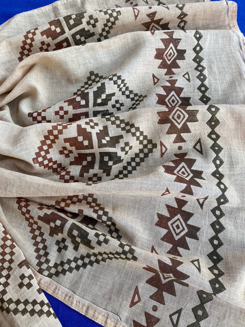 May include: A beige cotton scarf with a brown and black geometric pattern. The pattern is a repeating design of squares and triangles.