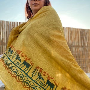 Peut inclure: &Eacute;charpe jaune moutarde avec un motif r&eacute;p&eacute;t&eacute; de cerfs et de formes g&eacute;om&eacute;triques en bleu sarcelle et rouille. L'&eacute;charpe est drap&eacute;e sur les &eacute;paules. Le fond est un ciel bleu clair.