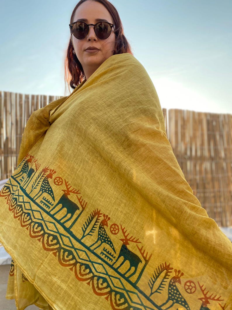 Peut inclure: &Eacute;charpe jaune moutarde avec un motif r&eacute;p&eacute;t&eacute; de cerfs et de formes g&eacute;om&eacute;triques en bleu sarcelle et rouille. L'&eacute;charpe est drap&eacute;e sur les &eacute;paules. Le fond est un ciel bleu clair.