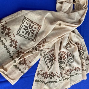 May include: A beige cotton scarf with a brown and green geometric pattern. The pattern is a repeating design of squares and triangles.