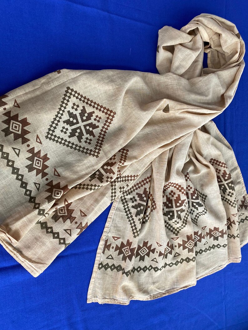 May include: A beige cotton scarf with a brown and green geometric pattern. The pattern is a repeating design of squares and triangles.
