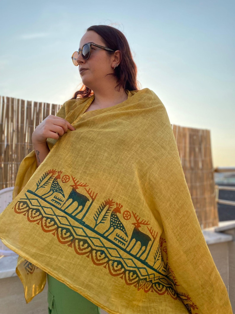 Peut inclure: &Eacute;charpe jaune moutarde avec une bordure d&eacute;corative repr&eacute;sentant des cerfs et des motifs g&eacute;om&eacute;triques en vert d'eau et rouille. L'&eacute;charpe est drap&eacute;e sur les &eacute;paules.