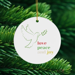 以下が含まれることがあります： 緑色のオリーブの枝と白い鳩が描かれた白いセラミック製のクリスマスオーナメント。オーナメントには「love peace and joy」という文字が赤、緑、黄色の色で書かれています。