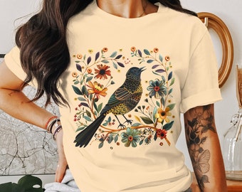 May include: A cream-colored t-shirt with a black bird and floral design. The bird is perched on a branch with colorful flowers and leaves.