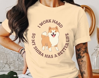 Op de afbeelding: Crèmekleurig T-shirt met een cartoon Shiba Inu-hond met een vrolijke uitdrukking. De tekst "I WORK HARD SO MY SHIBA HAS A BETTER LIFE" is rond de hond gedrukt. Het shirt heeft opgerolde mouwen.