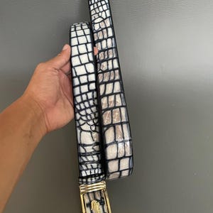 Op de afbeelding: Een wit en zwart riem met krokodillenpatroon en een gouden gesp. De riem wordt tegen een grijze achtergrond gehouden. De gesp heeft een gouden frame en een gedetailleerd embleem.