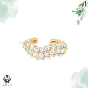 May include: A gold ear cuff featuring three rows of sparkling, round-cut white gemstones. The jewelry piece has a curved design and is set against a white background with subtle watercolor accents and the "ANCY JEWELRY" logo.