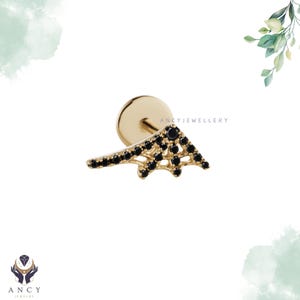 May include: A gold flat back stud earring featuring a triangular, web-like design adorned with small, round, black gemstones. The piece includes a circular backing and the text ANCYJEWELLERY printed in small, dark letters near the center.