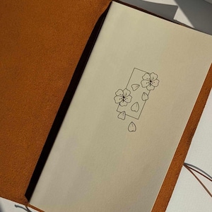 May include: A brown leather-bound journal with a blank page open. The page features a simple line drawing of a flower with petals falling from it.
