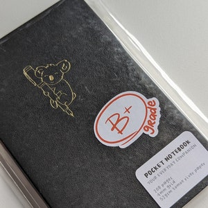 May include: A black leather-bound pocket notebook with a gold koala illustration on the cover. The notebook has a sticker on the front that says "B+ Grade" and includes the text "POCKET NOTEBOOK YOUR EVERYDAY COMPANION 208 pages 4mm grid 52gsm tomoe river paper".