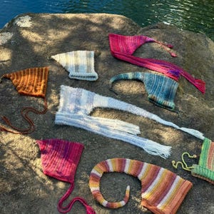 Può includere: Una collezione di cappelli colorati lavorati a mano in vari stili, tra cui cappelli da elfo a punta e modelli lunghi e fluenti. I cappelli presentano una gamma di colori, come arancione, rosa, blu e giallo, e sono esposti su una superficie di pietra strutturata.