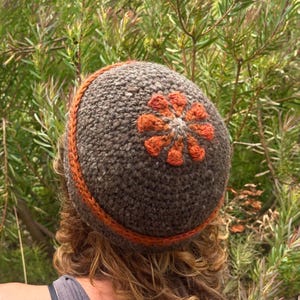 May include: A handmade, crocheted hat in shades of brown and orange. The hat features a textured, woolly appearance with an orange floral design on top and an orange band around the base. The hat is shown against a backdrop of green foliage.