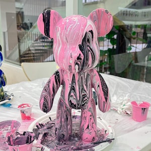 Può includere: Una statuetta di orsacchiotto di peluche marmoreggiata rosa e nera con finitura lucida. L'orso è in piedi su una superficie bianca con schizzi di vernice.