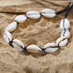 Könnte beinhalten: Eine weiße Muschelkette mit schwarzem Band. Die Kette besteht aus Kaurischnecken und ist auf einem schwarzen Band aufgefädelt.