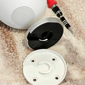 May include: A white security camera with a black base and a white mounting plate. The camera has a speaker grille and a small black button on the side. The base has a black ring around the edge and a small black button on the bottom. The mounting plate has four screw holes and a small white ring in the center.