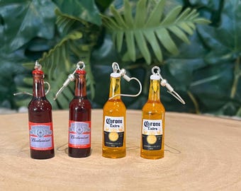 May include: Four pairs of beer bottle earrings. Two pairs are red and say "Budweiser" on the label. Two pairs are yellow and say "Corona Extra" on the label.
