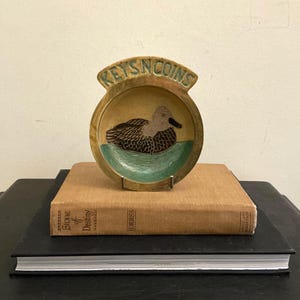 May include: A wooden dish with a duck motif and the text "KEYS N COINS". The dish is circular with a raised edge. The duck is brown and black, set against green water. The dish is displayed on a stack of books.