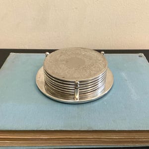 May include: A stack of circular silver-toned metal coasters held within a matching silver-toned metal holder. The top coaster features an intricate, embossed floral pattern surrounding a smooth, plain circular centre. The set is metallic.