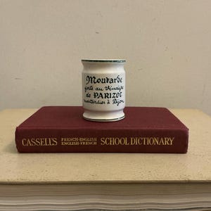 May include: A white ceramic mustard pot with green trim and black text, placed on a maroon book. The text on the pot reads "Moutarde forte au Vinaigre de PARIZOT moutardier à Dijon". The book is a dictionary.