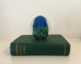 May include: An oval glass paperweight featuring a blue sky and landscape design. The paperweight rests on a dark green book. The book's spine displays the text "COLERIDGE'S POETICAL WORKS".