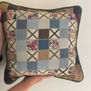 May include: Square cushion with a patchwork design. The cushion features a central grid of squares in shades of blue, brown, and beige, with floral accents. The border is a darker brown with a green trim.