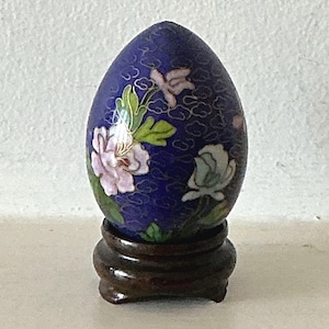 May include: A blue cloisonné egg with floral designs in pink, white, and green on a wooden stand.