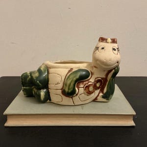 May include: A ceramic turtle-shaped planter in cream, green, and brown. The turtle has a smiling face and is resting on a light green book. The planter is designed with a hollow center for plants.